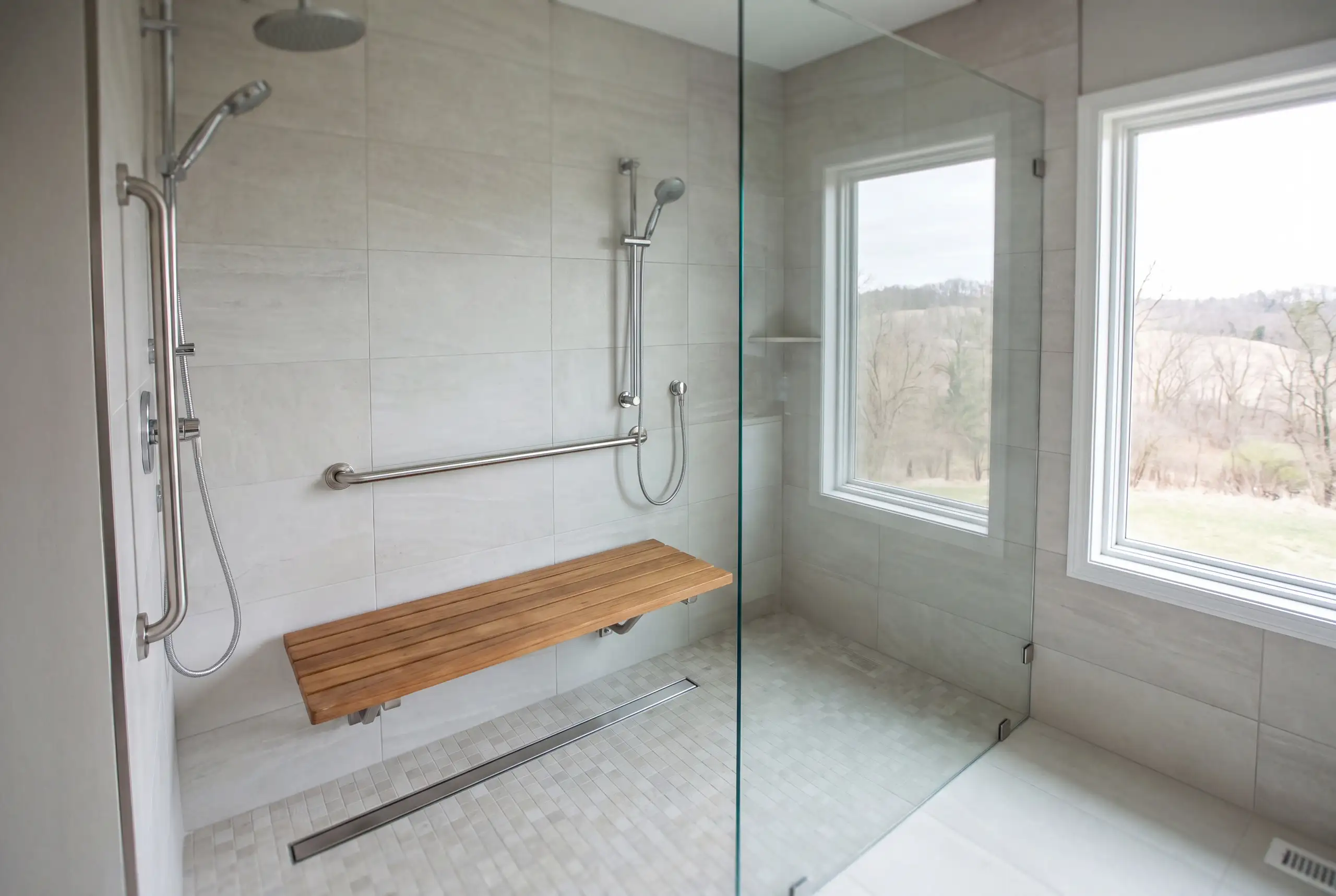 A curbless roll in shower with a folding teak bench, grab bars and a linear drain