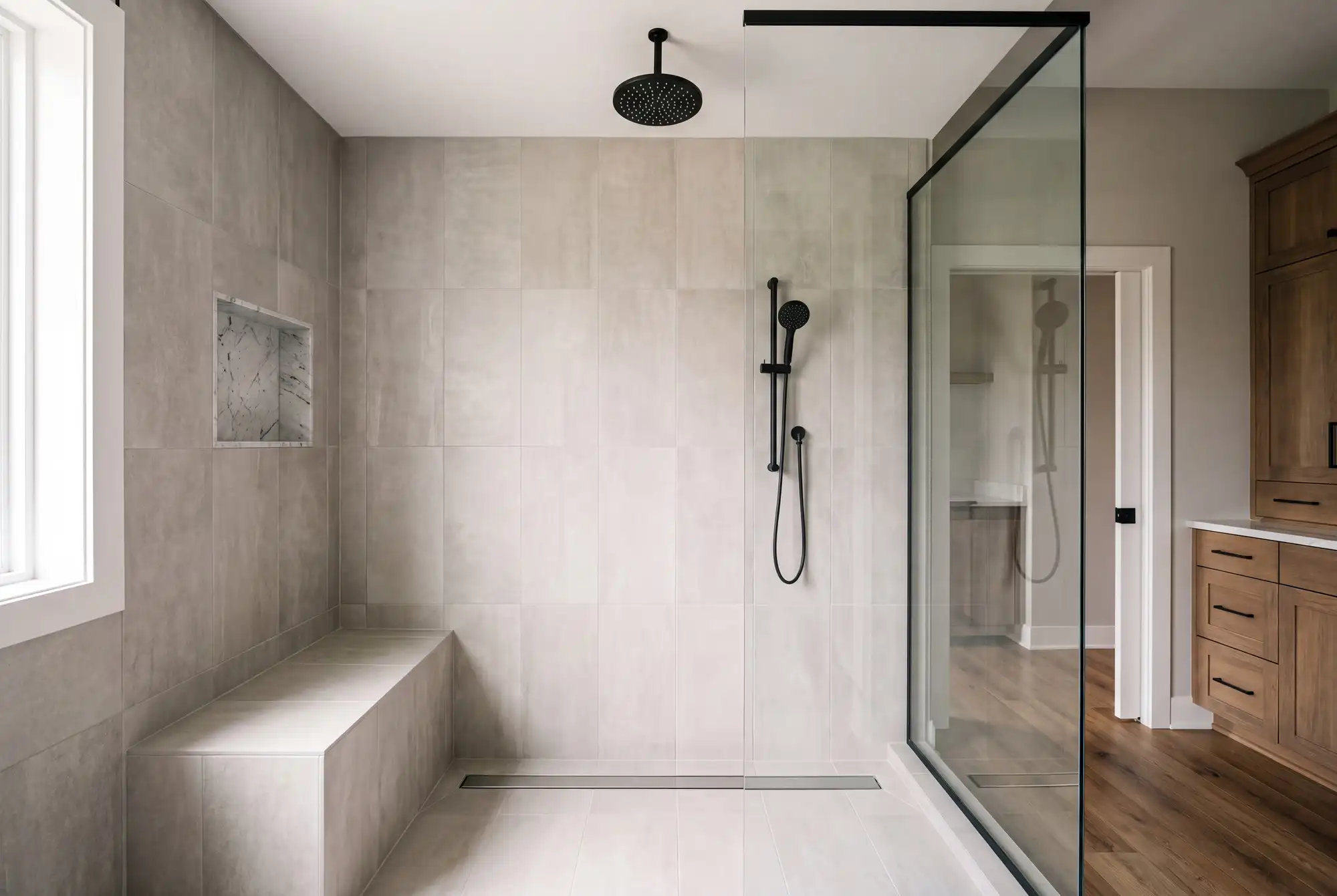 A walk in tile shower with a matte black rain head, a bench and a frameless glass panel